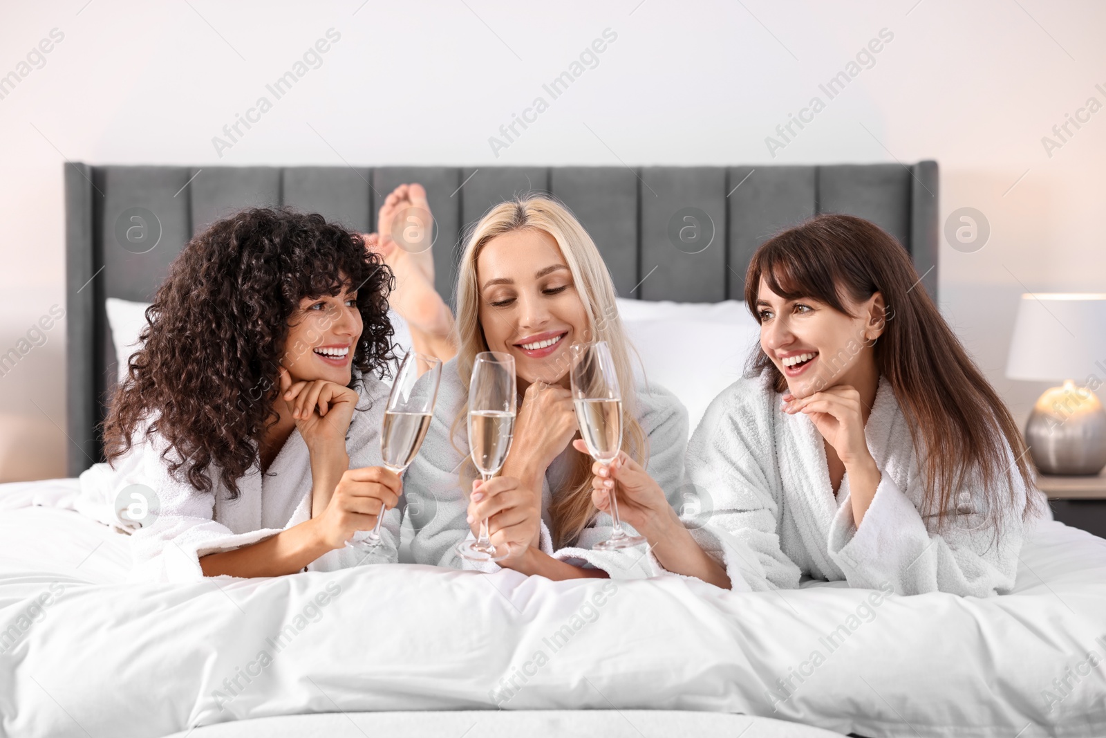 Photo of Happy friends with glasses of sparkling wine on bed. Spa party