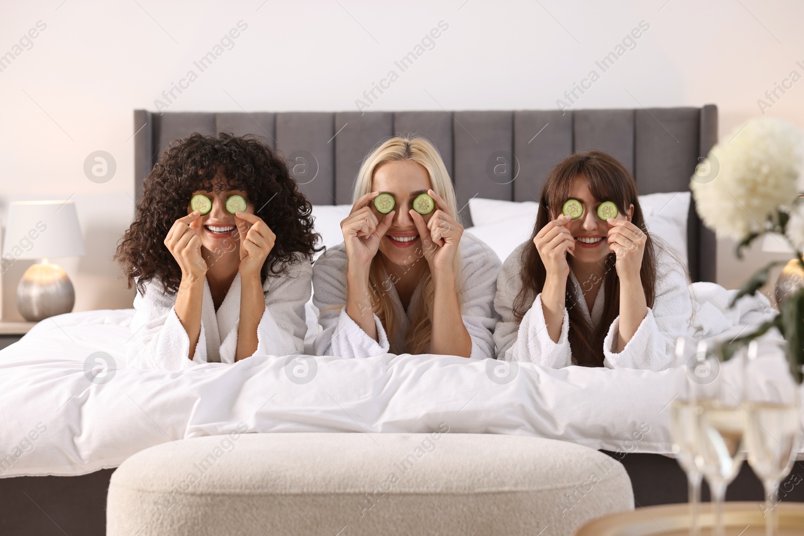 Photo of Happy friends with cucumber slices on bed. Spa party