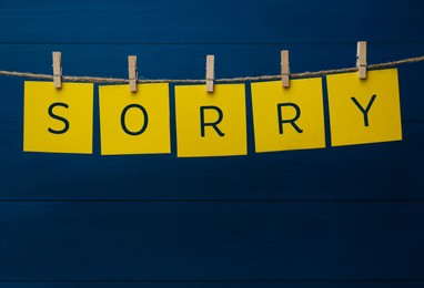 Word Sorry made of yellow paper notes with letters hanging on rope against blue wooden background