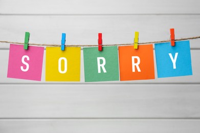 Word Sorry made of paper notes with letters hanging on rope against white wooden background