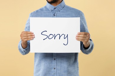 Man holding sheet of paper with word Sorry on beige background, closeup