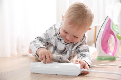 Photo of Little child playing with power strip and iron plug on floor indoors. Dangerous situation