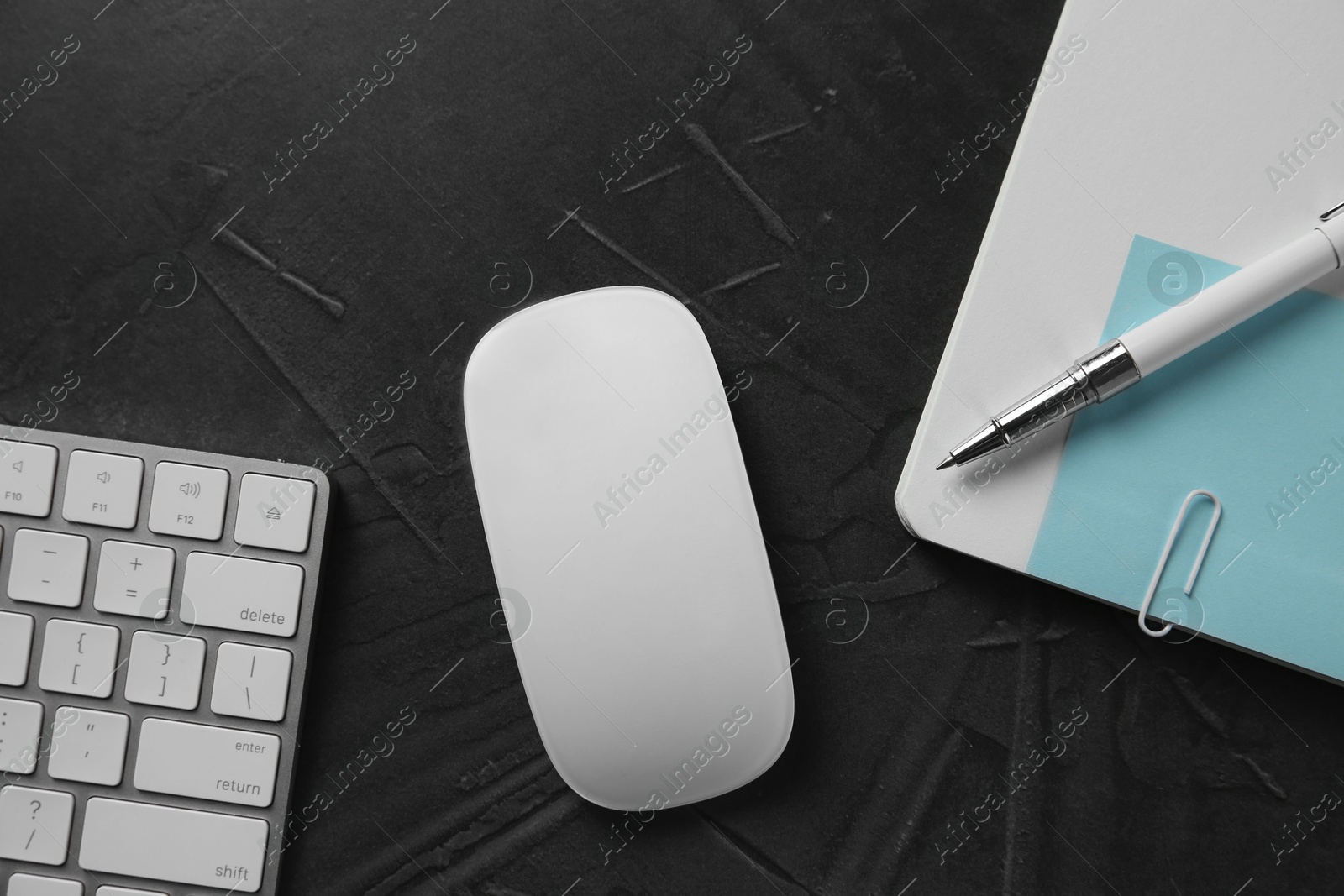 Photo of Wireless mouse, computer keyboard, notebook and pen on dark textured table, flat lay