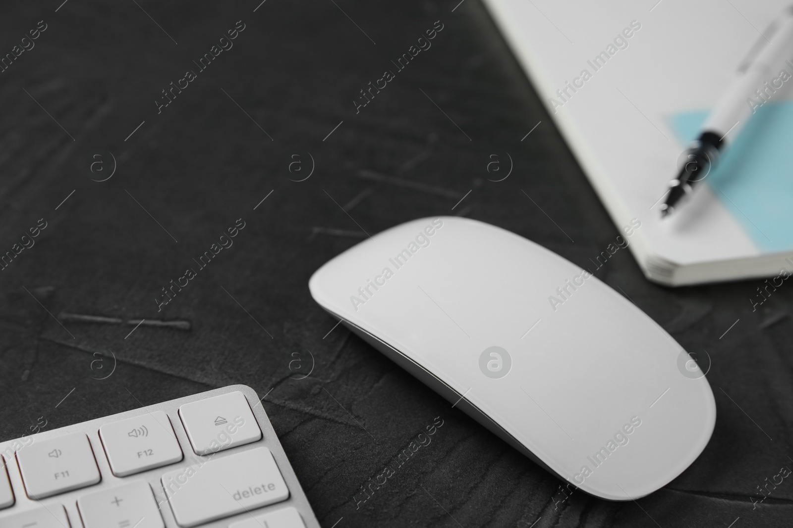 Photo of Wireless mouse, computer keyboard, notebook and pen on dark textured table, closeup
