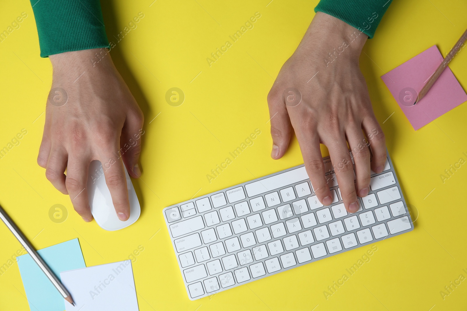 Photo of Man working with mouse, computer keyboard and stationery at yellow table, top view