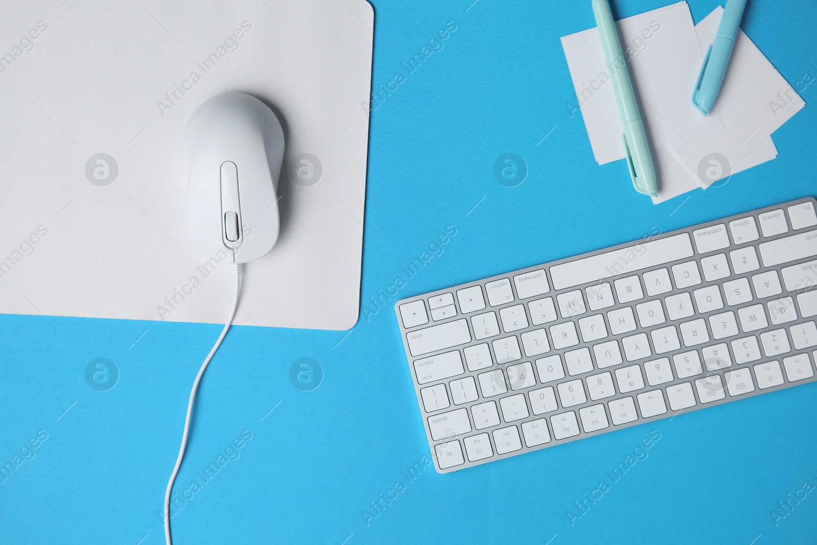 Photo of Wired mouse with mousepad, stationery and computer keyboard on light blue background, flat lay