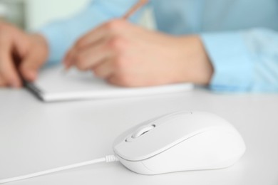 Wired mouse on white table, closeup. Man writing in notebook indoors, selective focus