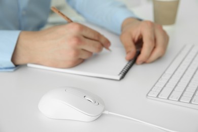 Wired mouse on white table, closeup. Man writing in notebook indoors, selective focus