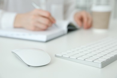 Photo of Wireless mouse on white table, closeup. Woman writing in notebook indoors, selective focus