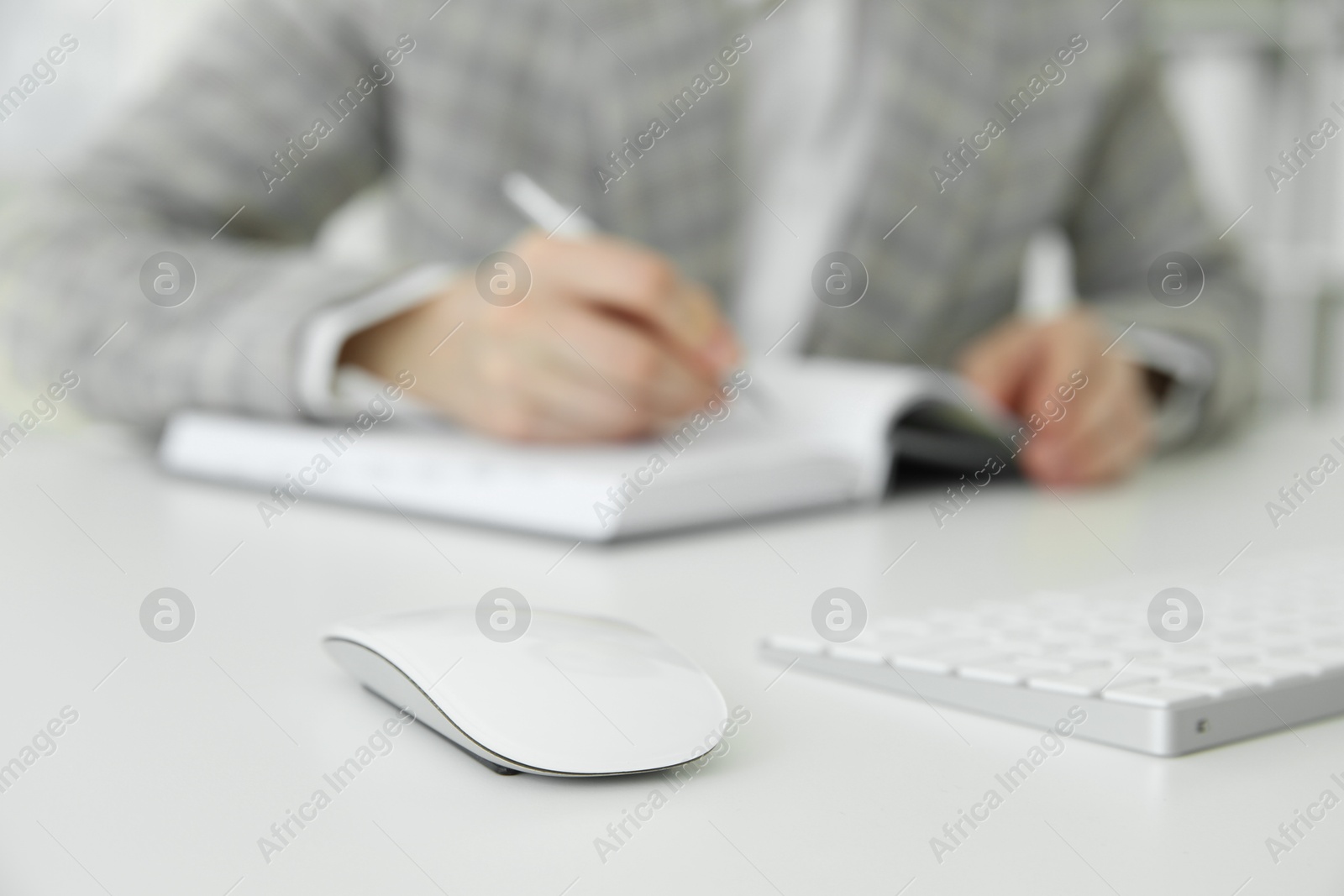 Photo of Wireless mouse on white table, closeup. Woman writing in notebook indoors, selective focus