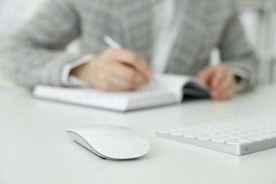 Photo of Wireless mouse on white table, closeup. Woman writing in notebook indoors, selective focus