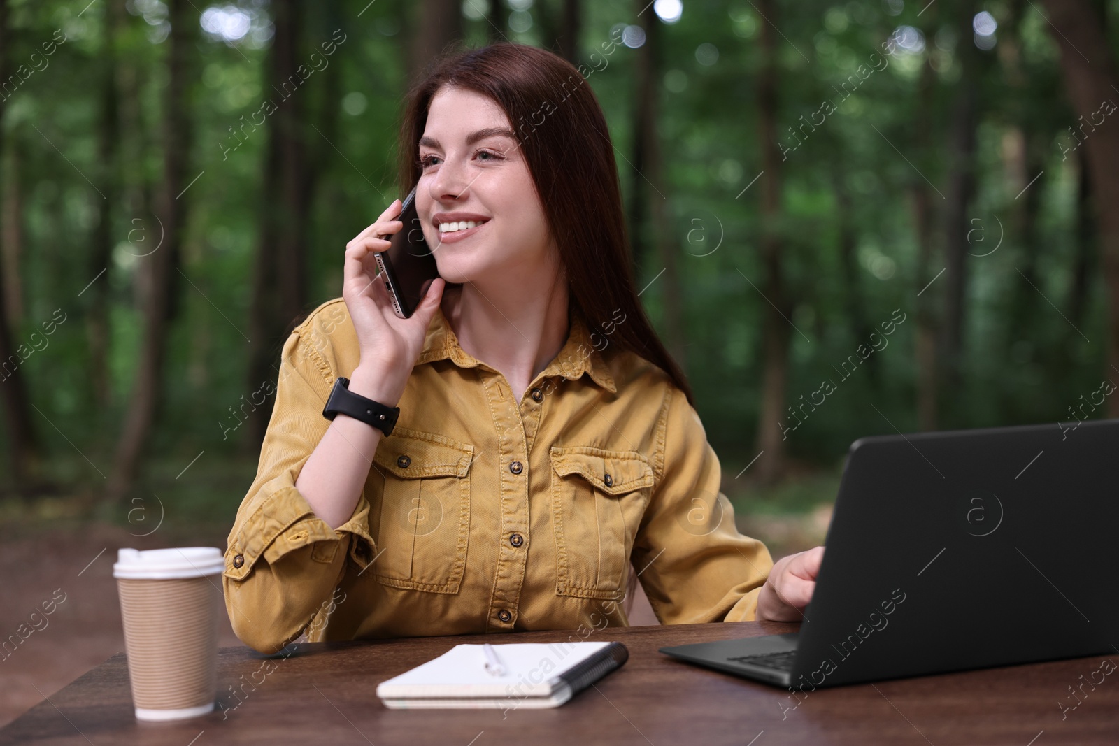 Photo of Smiling freelancer talking on smartphone at table with laptop outdoors. Remote job