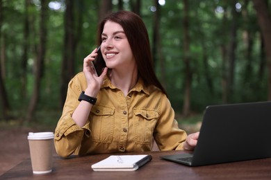 Photo of Smiling freelancer talking on smartphone at table with laptop outdoors. Remote job
