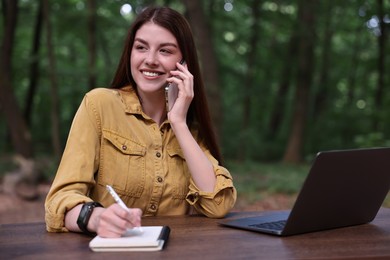 Smiling freelancer talking on smartphone at table with laptop outdoors. Remote job