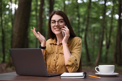 Photo of Smiling freelancer talking on smartphone at table with laptop outdoors. Remote job