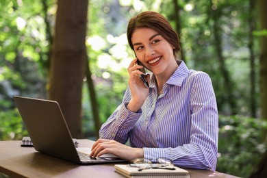 Smiling businesswoman talking on smartphone at table with laptop outdoors. Remote job