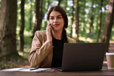Photo of Businesswoman talking on smartphone at table with laptop outdoors. Remote job