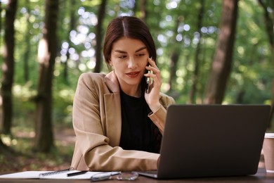 Businesswoman talking on smartphone at table with laptop outdoors. Remote job