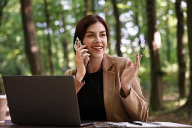 Smiling businesswoman talking on smartphone at table with laptop outdoors. Remote job