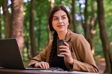 Photo of Businesswoman with paper cup working on laptop at table outdoors. Remote job