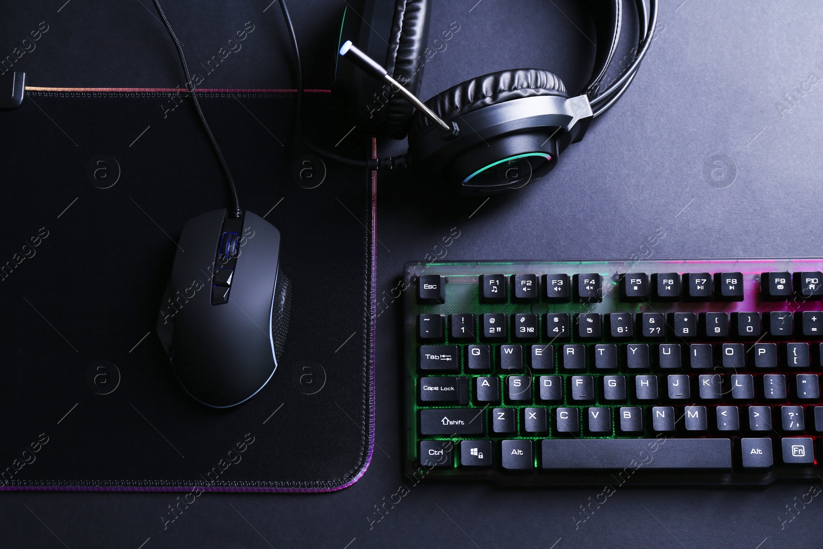 Photo of Wired mouse, mousepad, computer keyboard and headset on black table, flat lay