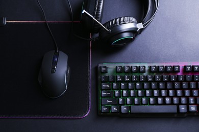 Photo of Wired mouse, mousepad, computer keyboard and headset on black table, flat lay