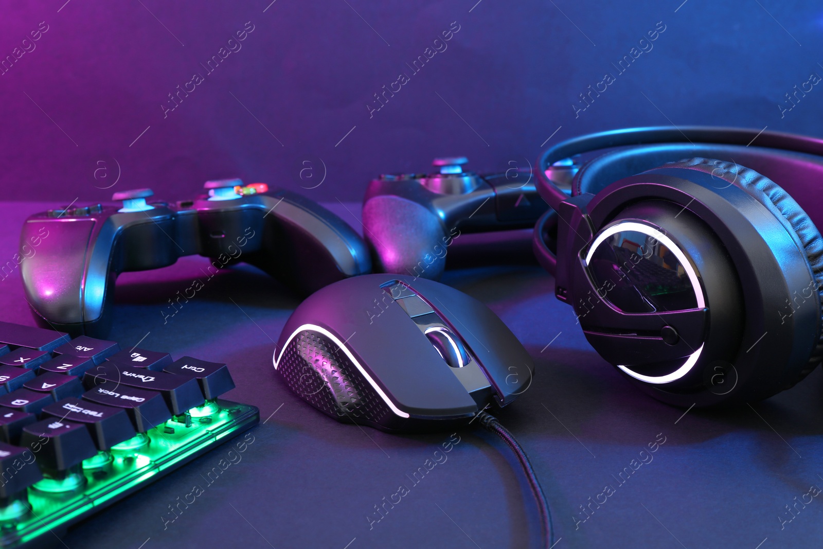 Photo of Computer mouse, headset, RGB keyboard and game controllers in neon lights on dark background, closeup