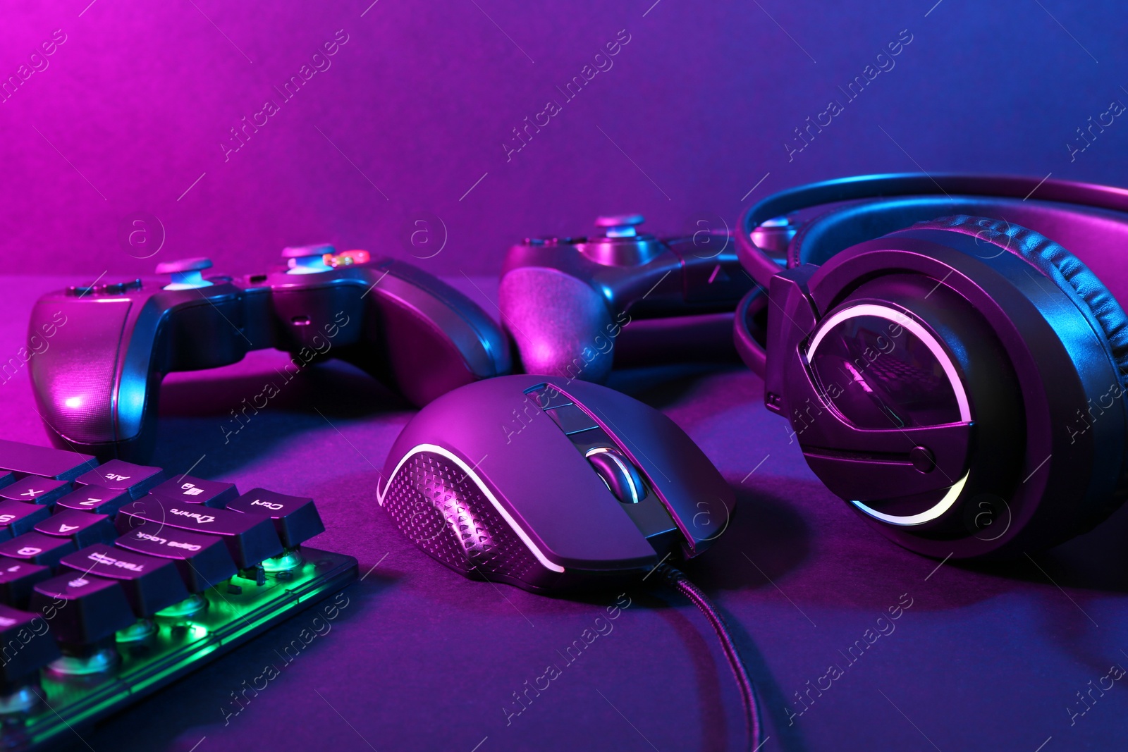 Photo of Computer mouse, headset, RGB keyboard and game controllers in neon lights on dark background, closeup