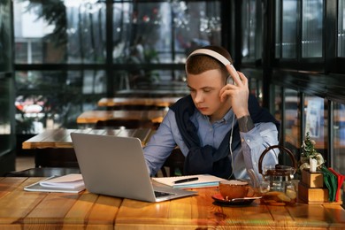 Young male student with laptop and headphones studying at table in cafe