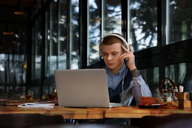 Young male student with laptop and headphones studying at table in cafe