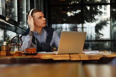 Young male student with laptop and headphones studying at table in cafe