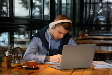 Young male student with laptop and headphones studying at table in cafe
