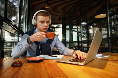 Young male student with laptop drinking coffee while studying at table in cafe