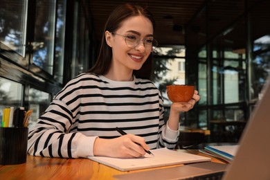 Photo of Young female student with laptop drinking coffee while studying at table in cafe