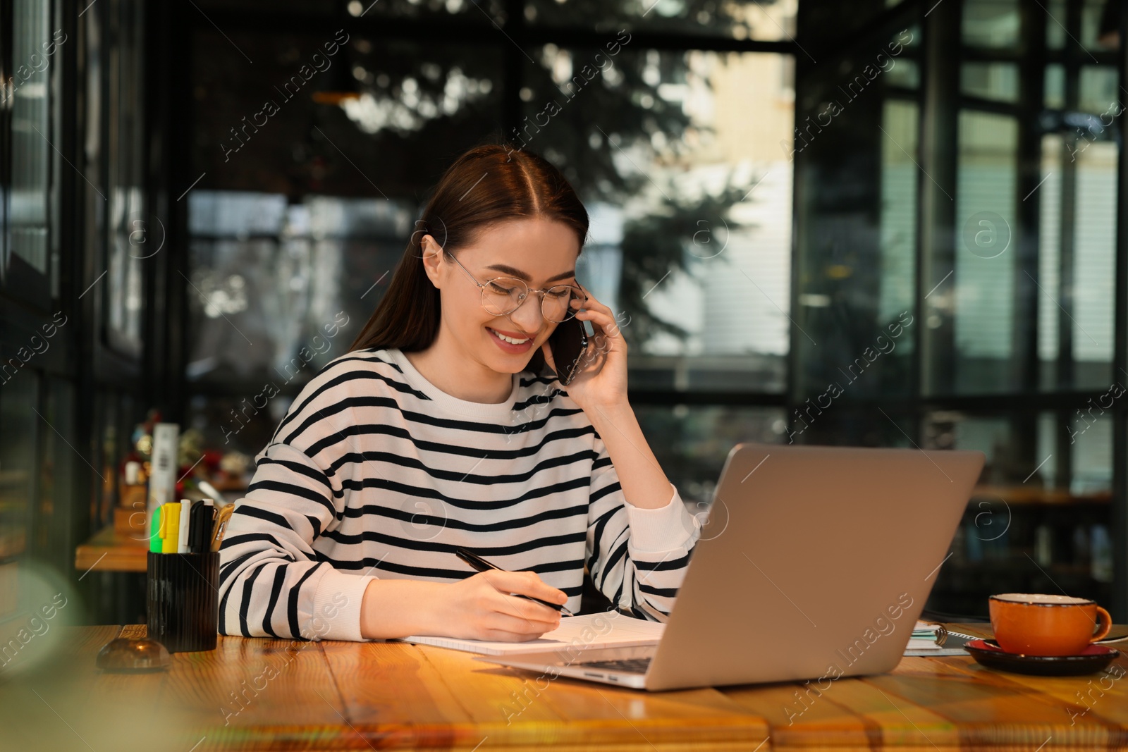 Photo of Young female student with laptop talking on phone while studying at table in cafe