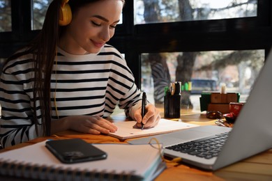 Photo of Young female student with laptop and headphones studying at table in cafe