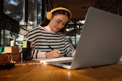 Young female student with laptop and headphones studying at table in cafe