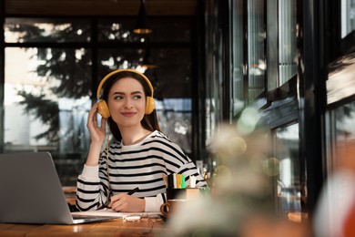 Young female student with laptop and headphones studying at table in cafe