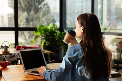 Young female student with laptop drinking coffee while studying at table in cafe