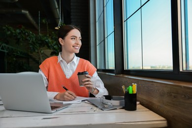 Young female student with laptop drinking coffee while studying at table in cafe