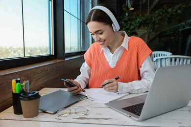 Young female student with laptop using smartphone while studying at table in cafe