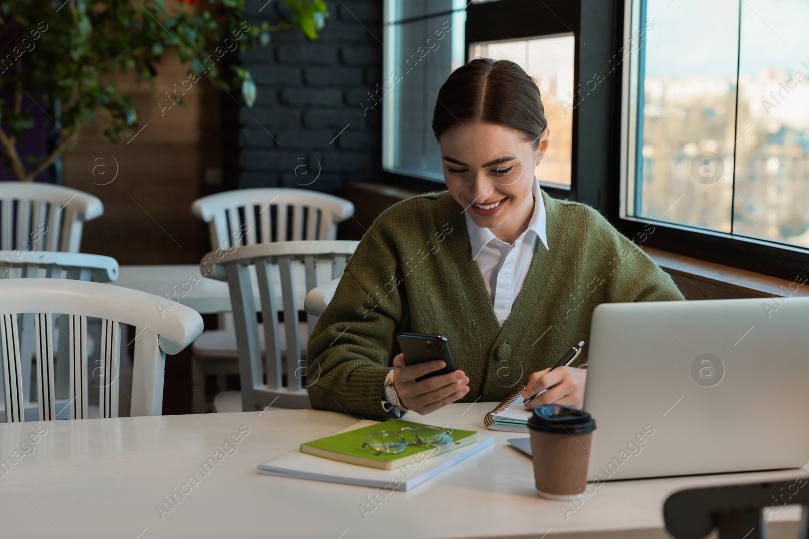 Photo of Young female student with laptop using smartphone while studying at table in cafe