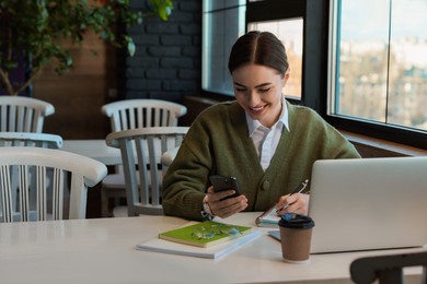 Young female student with laptop using smartphone while studying at table in cafe