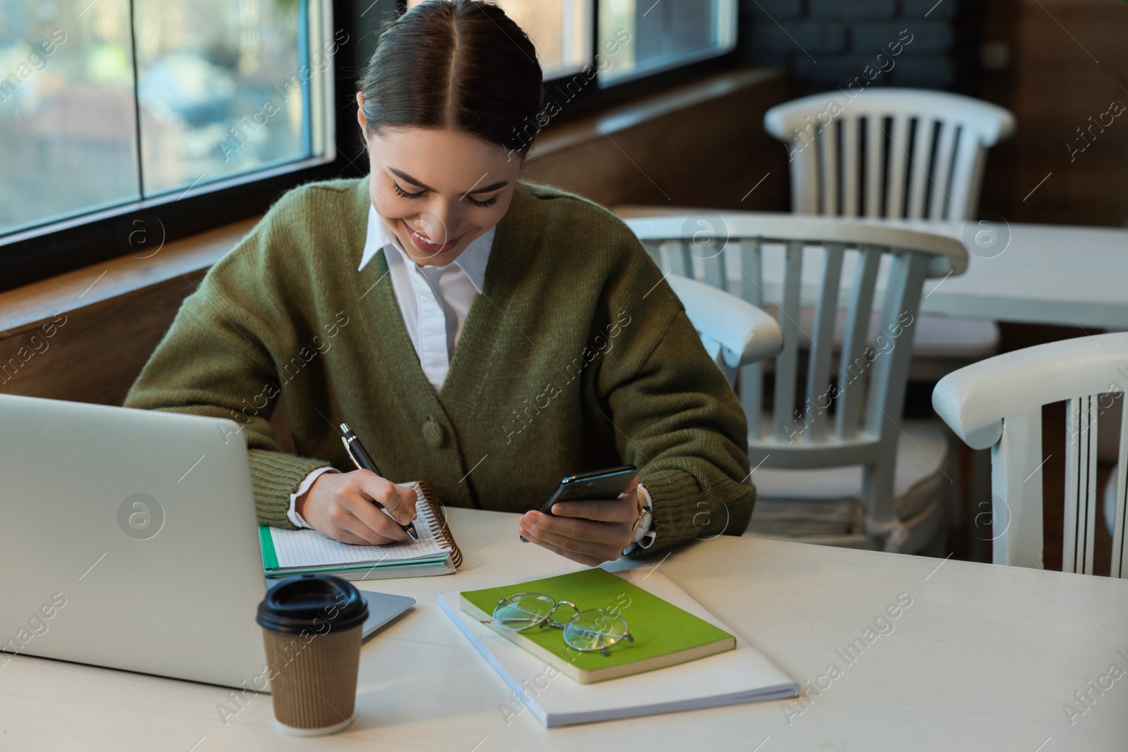 Photo of Young female student with laptop using smartphone while studying at table in cafe