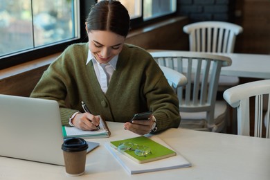 Photo of Young female student with laptop using smartphone while studying at table in cafe