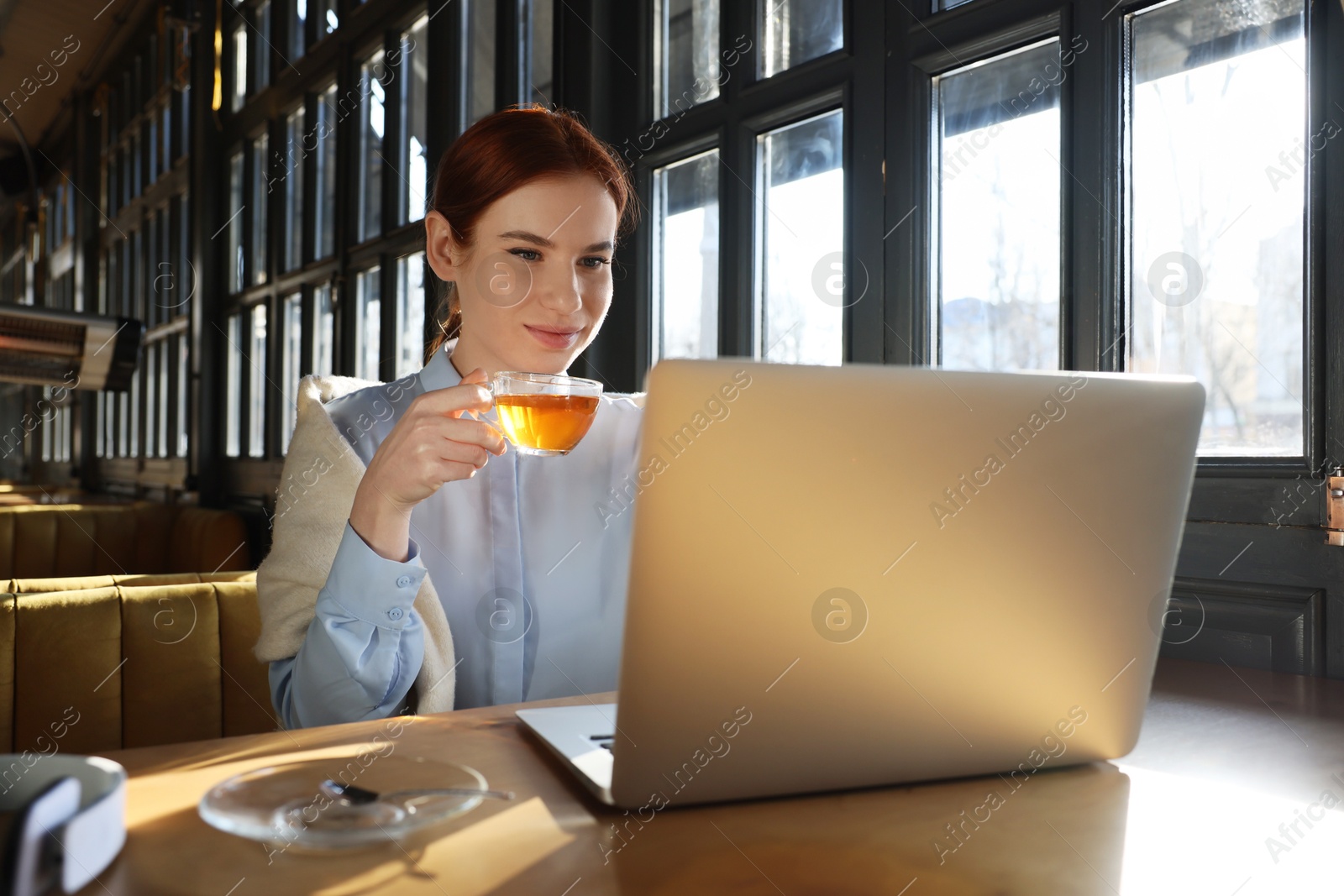 Photo of Young female student with laptop drinking tea while studying at table in cafe