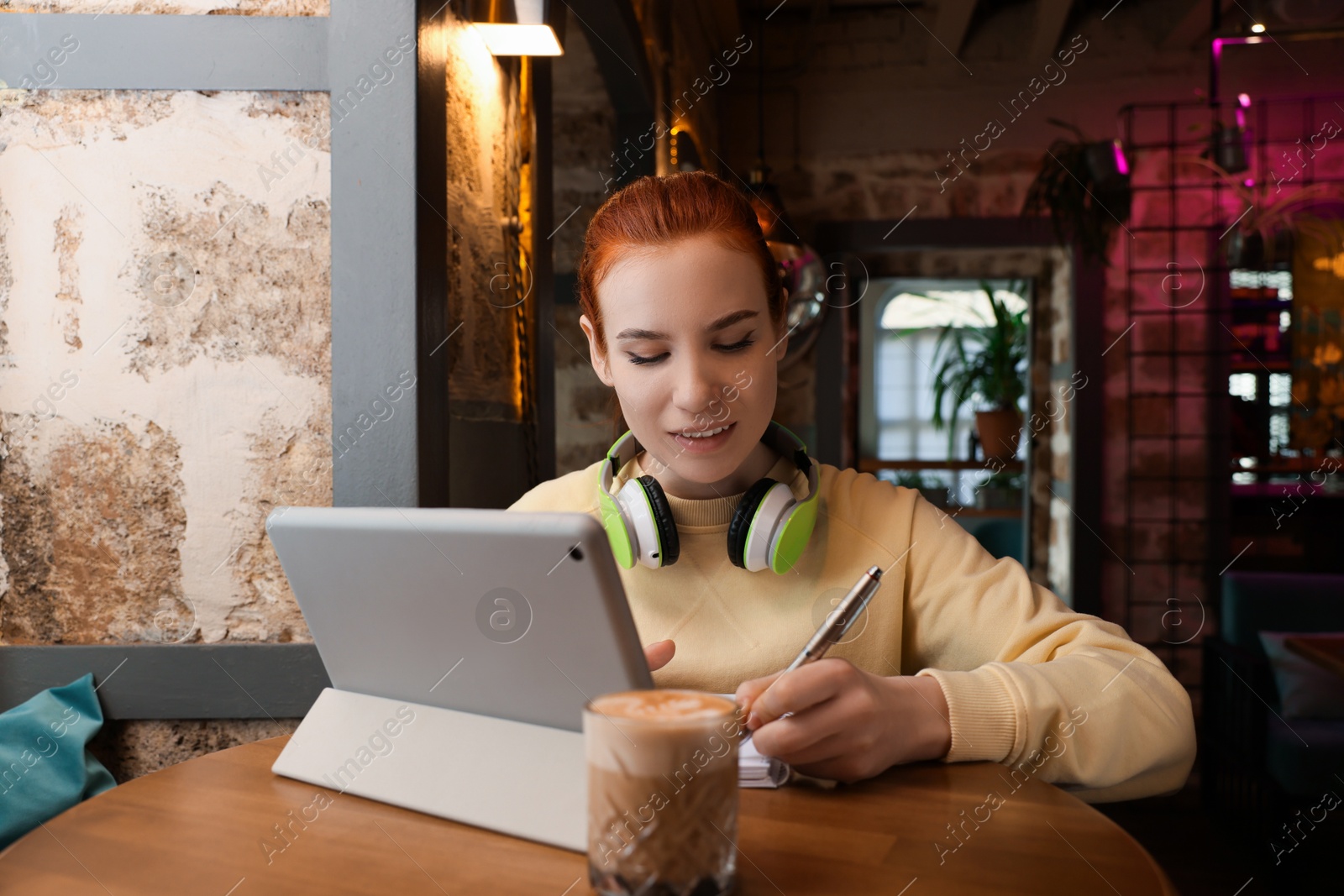 Photo of Young female student with tablet and headphones studying at table in cafe