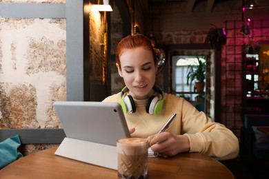 Photo of Young female student with tablet and headphones studying at table in cafe