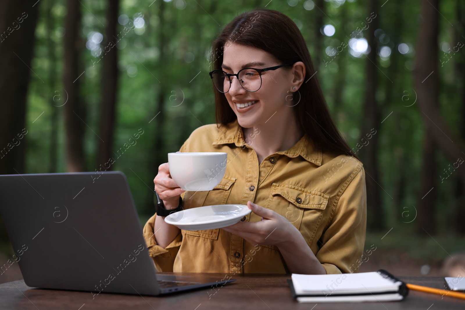 Photo of Smiling freelancer drinking coffee at table with laptop outdoors. Remote job