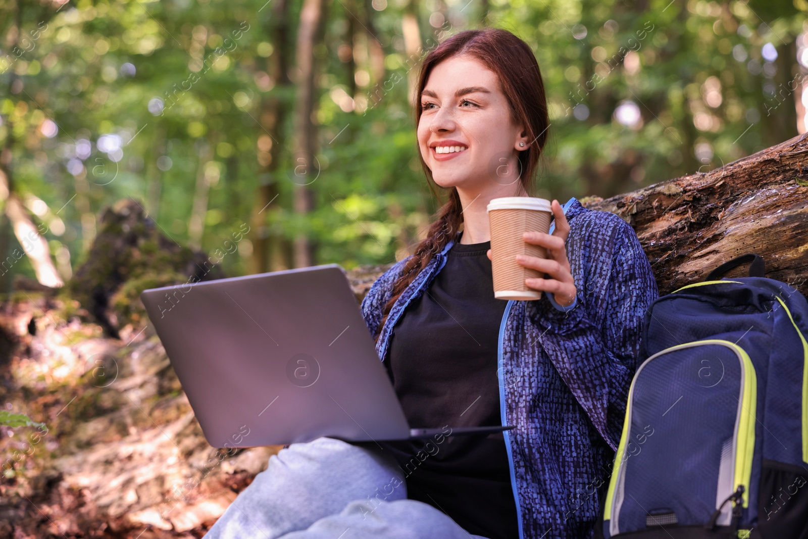 Photo of Smiling freelancer drinking coffee with laptop in forest, low angle view. Remote work